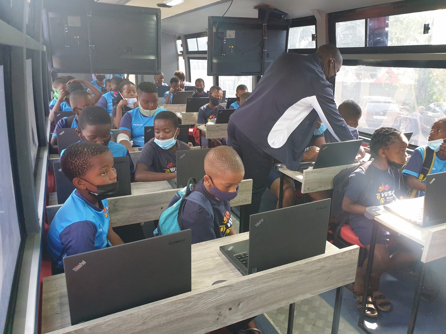 Learners working at laptops inside the DigiBus while a facilitator helps a pupil at his desk